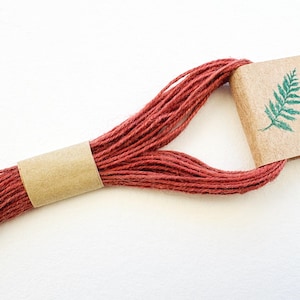 May include: A skein of deep terracotta red embroidery floss secured by a plain tan paper band and a tan paper label featuring a small green fern leaf illustration. The thread is looped and held together by the paper components.