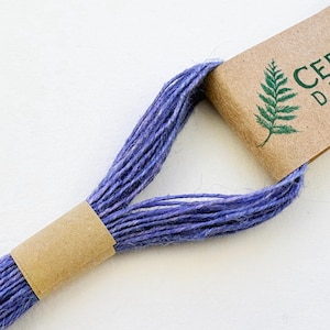 May include: A skein of purple embroidery thread secured by two brown paper bands. The top band features a green fern illustration and partial text reading CE and D. The thread has a textured, fibrous appearance with visible individual strands.