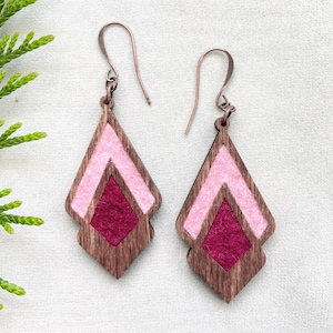 Könnte beinhalten: Ein Paar Holzohrringe mit rosa und burgunderfarbenen Filz-Akzenten. Die Ohrringe sind dreieckig mit einem kleineren Dreieck im Inneren.
