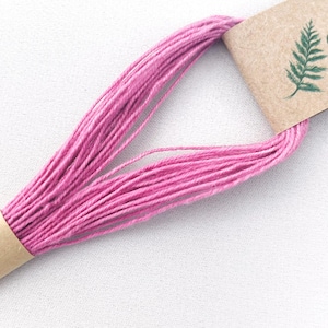 May include: A bundle of vibrant pink embroidery thread looped and secured by two brown paper bands. One band features a small, detailed green fern leaf illustration printed on its surface, providing a natural accent to the bright pink fibre.