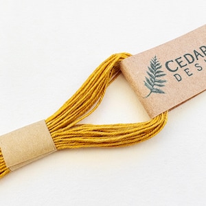 May include: A skein of mustard yellow embroidery thread looped through a brown paper label. The label features a dark green fern illustration and the partial text CEDAR DESI printed in a dark font on the textured tan paper surface.