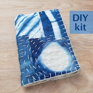 Op de afbeelding: Een rechthoekige stoffen kaft met een blauw en wit tie-dye patroon met zichtbare witte rijgsteken in sashiko-stijl. De kaft toont een cirkelvormig motief en gestreepte secties. Een blauwe rechthoekige afbeelding met de tekst DIY kit is zichtbaar.