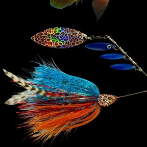 May include: A pumpkinseed sunfish with orange and blue speckled scales positioned above a fishing spinner bait. The lure features a holographic leopard print blade, three small blue blades, and a skirt with blue, orange, and white feathers.