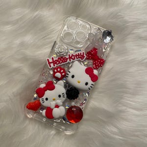 May include: Clear phone case with a Hello Kitty design. The case features two Hello Kitty figures, red bows, glitter, and rhinestones.