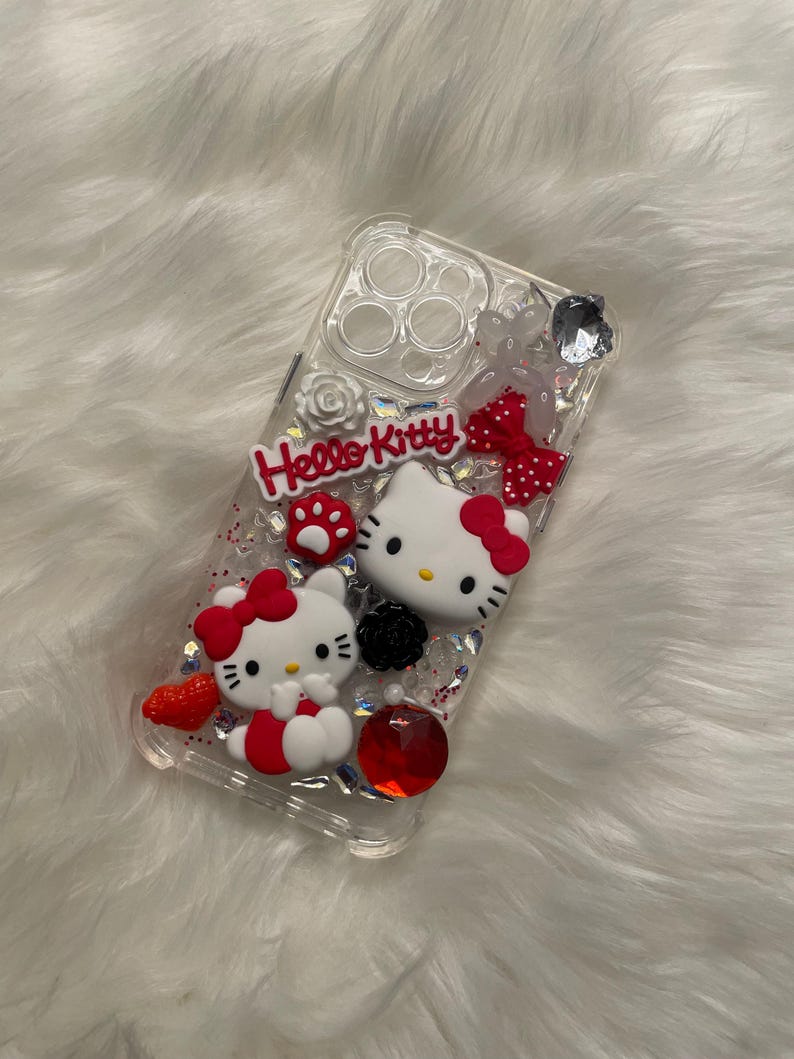 May include: Clear phone case with a Hello Kitty design. The case features two Hello Kitty figures, red bows, glitter, and rhinestones.