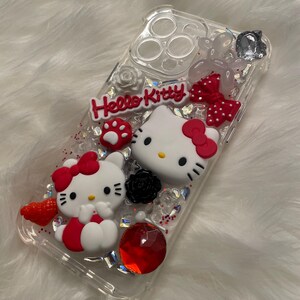 May include: Clear phone case with Hello Kitty design. The case features two Hello Kitty figures, one with a red bow and the other with a pink bow. The case is decorated with glitter, rhinestones, and other embellishments.