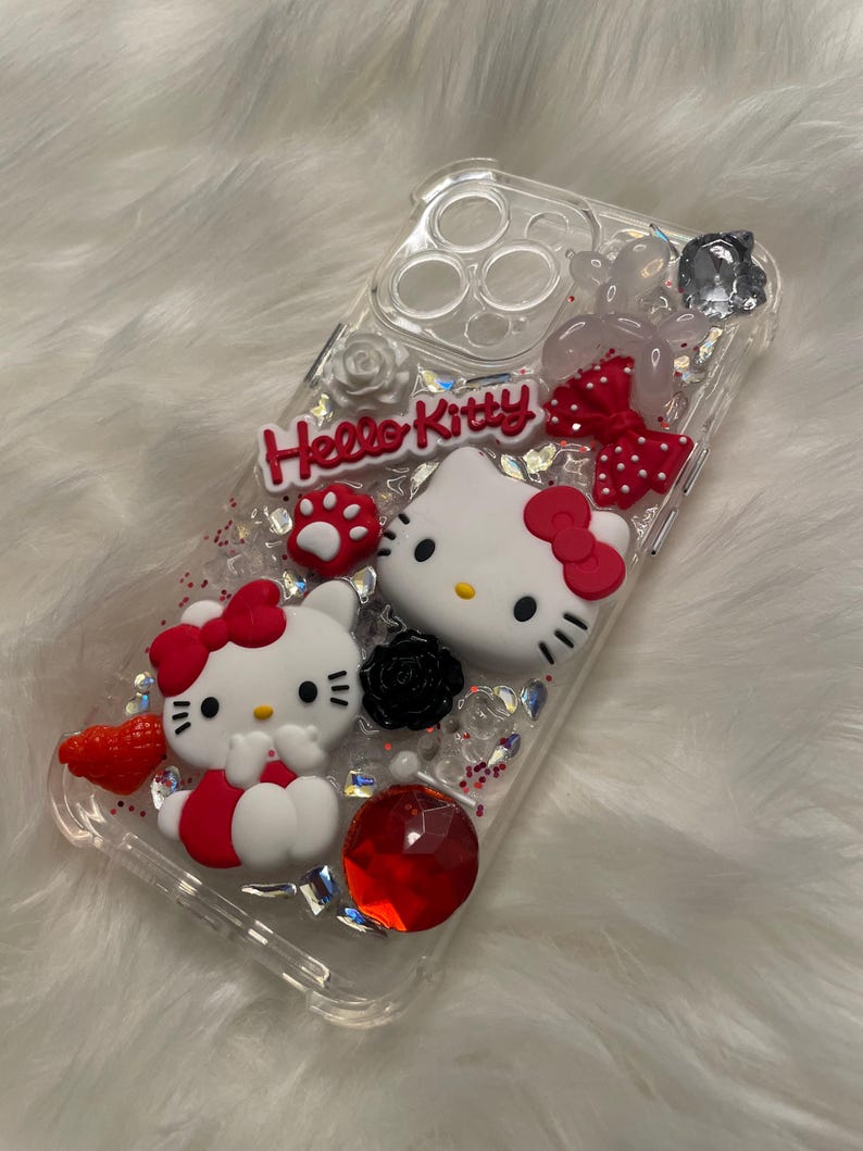 May include: Clear phone case with Hello Kitty design. The case features two Hello Kitty figures, one with a red bow and the other with a pink bow. The case is decorated with glitter, rhinestones, and other embellishments.