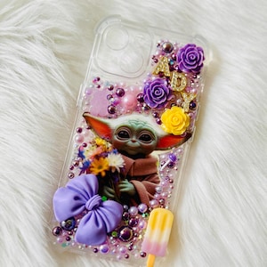 Op de afbeelding: Een transparante telefoonhoes met een paarse strik, bloemen en een afbeelding van Baby Yoda die bloemen vasthoudt. De hoes heeft ook gouden letters die "ADY" spellen en een roze en gele ijslolly.