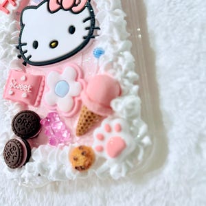 Pu&ograve; includere: Una custodia per telefono rosa Hello Kitty con glassa bianca e varie decorazioni, tra cui un cono gelato rosa, un fiore, un'impronta di zampa e biscotti Oreo.