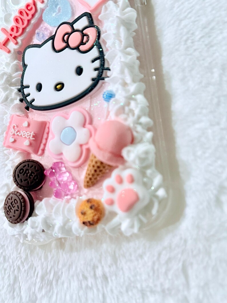 Pu&ograve; includere: Una custodia per telefono rosa Hello Kitty con glassa bianca e varie decorazioni, tra cui un cono gelato rosa, un fiore, un'impronta di zampa e biscotti Oreo.