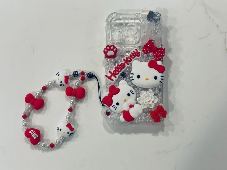 Puede incluir: Una funda transparente para tel&eacute;fono con un dise&ntilde;o de Hello Kitty. La funda est&aacute; decorada con pedrer&iacute;a, lazos y personajes de Hello Kitty. Se incluye una correa para tel&eacute;fono de cuentas blancas y rojas con abalorios de Hello Kitty.