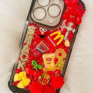 Op de afbeelding: Een transparante telefoonhoes met een rood, geel en zwart ontwerp. De hoes is versierd met verschillende voedselthema-charms, waaronder een McDonald's-logo, een zak Cheetos, een zak Lay's-chips en een kers. De hoes heeft ook een rode strik, een rode roos en een gouden kruis.