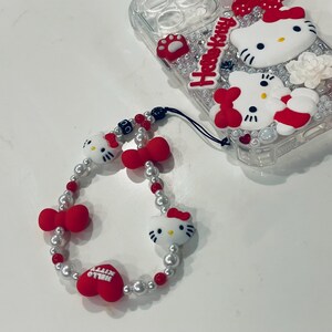 Puede incluir: Una funda para tel&eacute;fono Hello Kitty blanca y roja con la parte posterior transparente y un charm Hello Kitty a juego con una cadena de perlas y cuentas rojas.