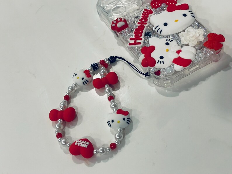 Puede incluir: Una funda para tel&eacute;fono Hello Kitty blanca y roja con la parte posterior transparente y un charm Hello Kitty a juego con una cadena de perlas y cuentas rojas.