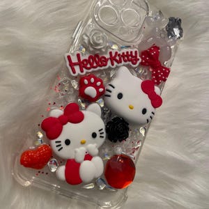 May include: Clear phone case with a red, white, and black Hello Kitty design. The case has glitter and rhinestones.