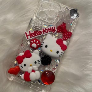 May include: Clear phone case with a red, white, and black Hello Kitty design. The case features two Hello Kitty characters, glitter, and rhinestones.