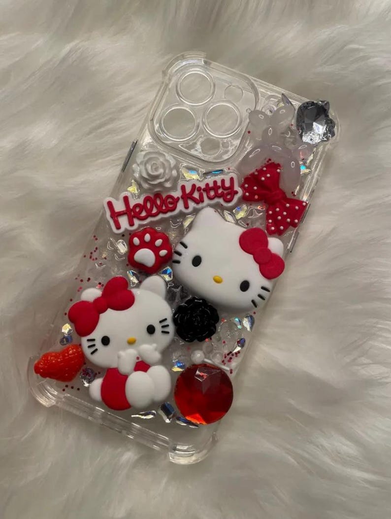 May include: Clear phone case with a red, white, and black Hello Kitty design. The case features two Hello Kitty characters, glitter, and rhinestones.