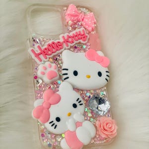 May include: A clear phone case with a pink Hello Kitty design. The case features two Hello Kitty characters, a pink bow, and glitter.