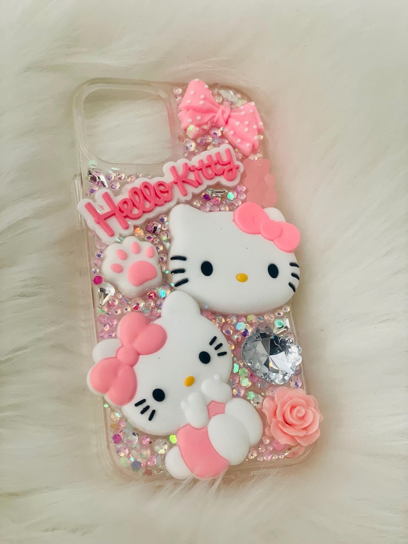 May include: A clear phone case with a pink Hello Kitty design. The case features two Hello Kitty characters, a pink bow, and glitter.