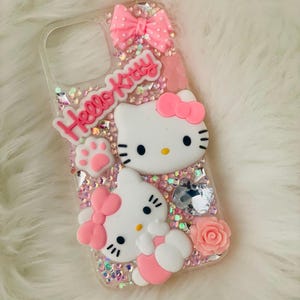 May include: A clear phone case with a pink Hello Kitty design. The case is decorated with glitter, rhinestones, and 3D Hello Kitty figures. The case has a pink bow and a pink paw print.