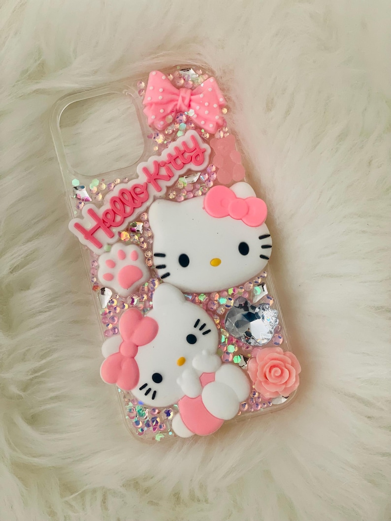 May include: A clear phone case with a pink Hello Kitty design. The case is decorated with glitter, rhinestones, and 3D Hello Kitty figures. The case has a pink bow and a pink paw print.