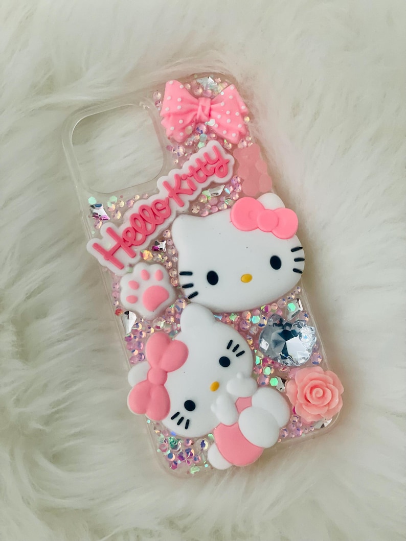 May include: A clear phone case with a pink Hello Kitty design. The case features two Hello Kitty characters, a pink bow, and glitter.