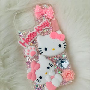 May include: A clear phone case with a pink Hello Kitty design. The case features two Hello Kitty figures, a pink bow, a pink paw print, and pink glitter.