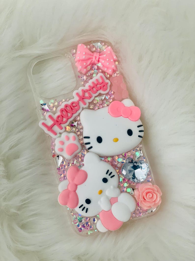 May include: A clear phone case with a pink Hello Kitty design. The case features two Hello Kitty figures, a pink bow, a pink paw print, and pink glitter.