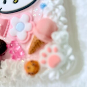 Pu&ograve; includere: Una custodia per telefono rosa e bianca con un design Hello Kitty, un fiore, un'impronta di zampa e un biscotto. La custodia &egrave; ricoperta di zuccherini bianchi.
