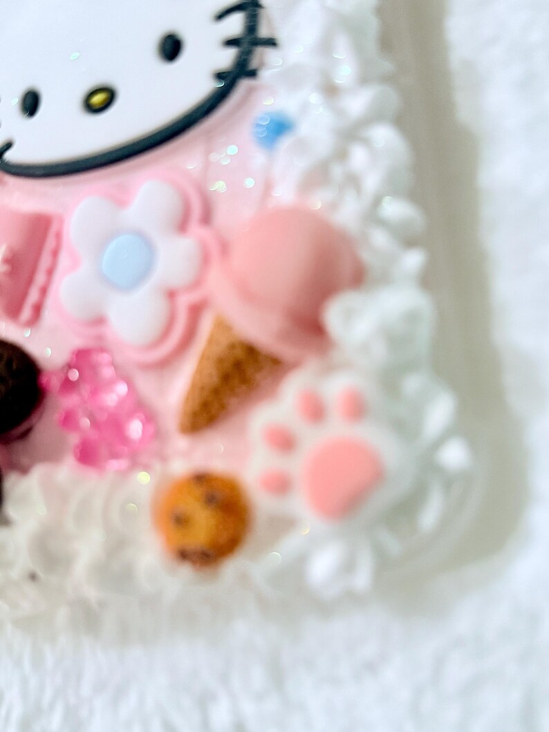 Pu&ograve; includere: Una custodia per telefono rosa e bianca con un design Hello Kitty, un fiore, un'impronta di zampa e un biscotto. La custodia &egrave; ricoperta di zuccherini bianchi.