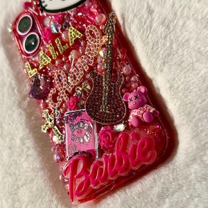 May include: A pink phone case decorated with rhinestones, glitter, and various charms, including a Hello Kitty character, a guitar, a teddy bear, and the word "Barbie".