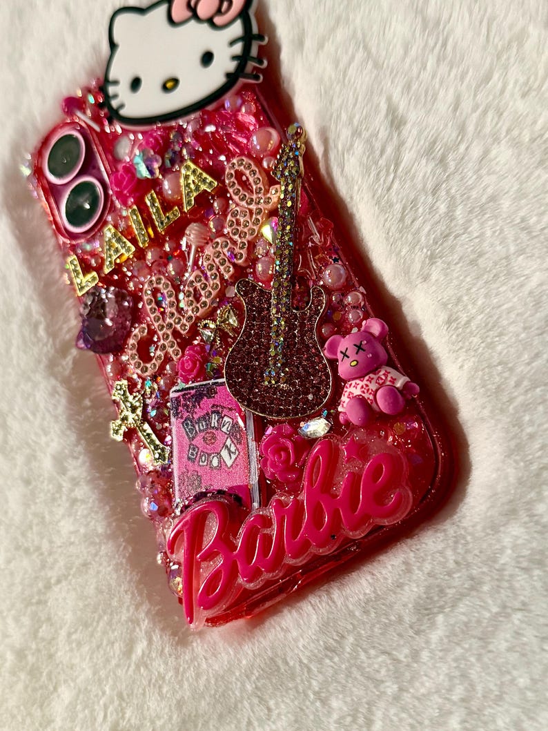 May include: A pink phone case decorated with rhinestones, glitter, and various charms, including a Hello Kitty character, a guitar, a teddy bear, and the word "Barbie".