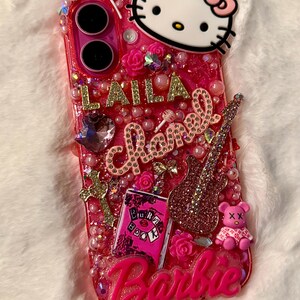 May include: A pink phone case with a Hello Kitty character, a pink guitar, a pink book with the words "Burn Book" on it, and the word "Barbie" in pink letters. The case is decorated with pink and gold glitter, rhinestones, and pearls.