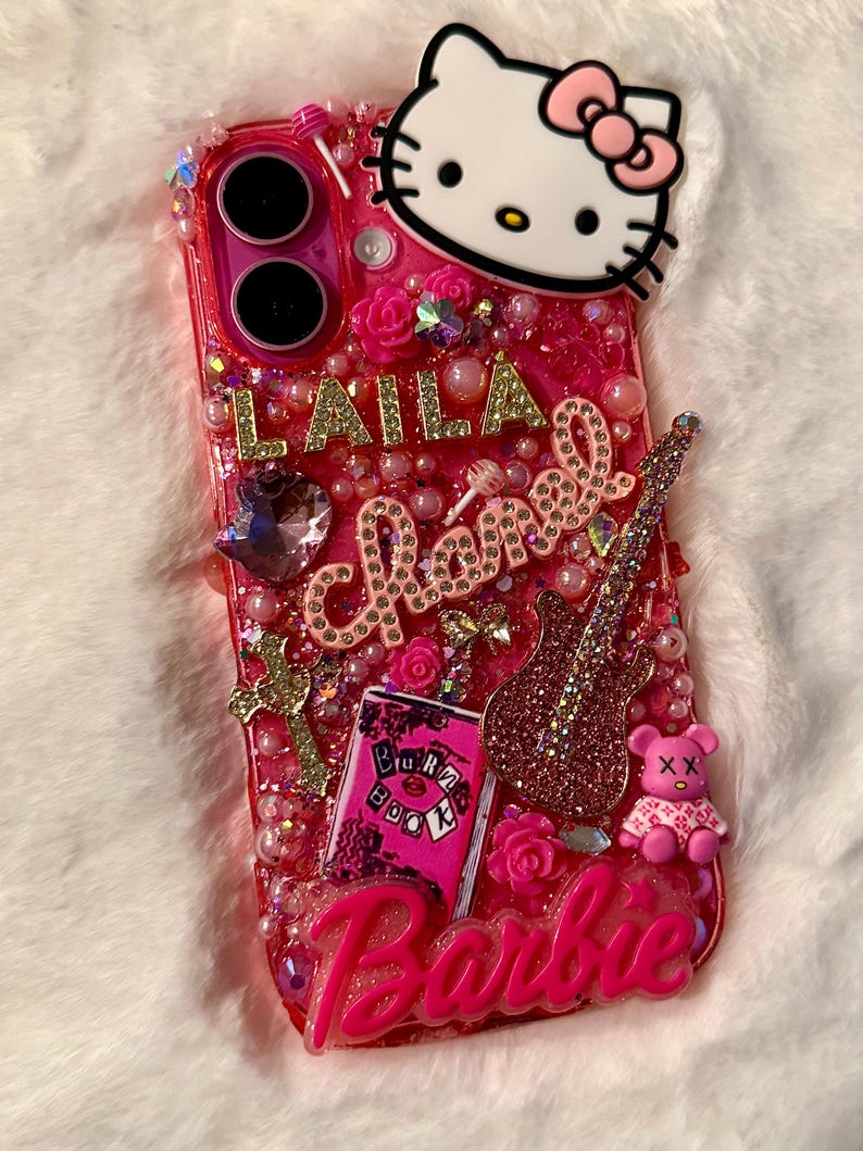 May include: A pink phone case featuring a Hello Kitty character, a pink guitar, a pink book with the words "Burn Book" on it, and the word "Barbie" in pink letters. The case is decorated with pink and gold glitter, rhinestones, and pearls.