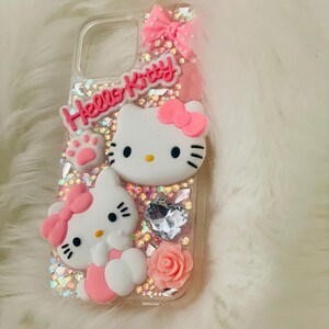 May include: A clear phone case with a pink Hello Kitty design. The case features two Hello Kitty figures, a pink bow, a pink rose, and glitter.