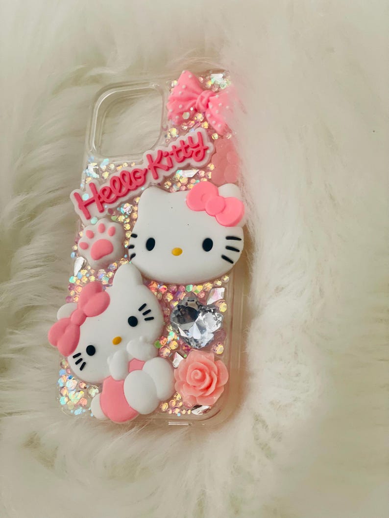 May include: A clear phone case with a pink Hello Kitty design. The case features two Hello Kitty figures, a pink bow, a pink rose, and glitter.