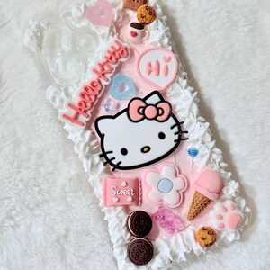 Pu&ograve; includere: Una custodia per telefono rosa e bianca Hello Kitty con un design Hello Kitty 3D, un cuore rosa, una tazza di caff&egrave;, un biscotto, un lecca-lecca, un fiore, un cono gelato e la parola "Ciao" in un fumetto rosa.