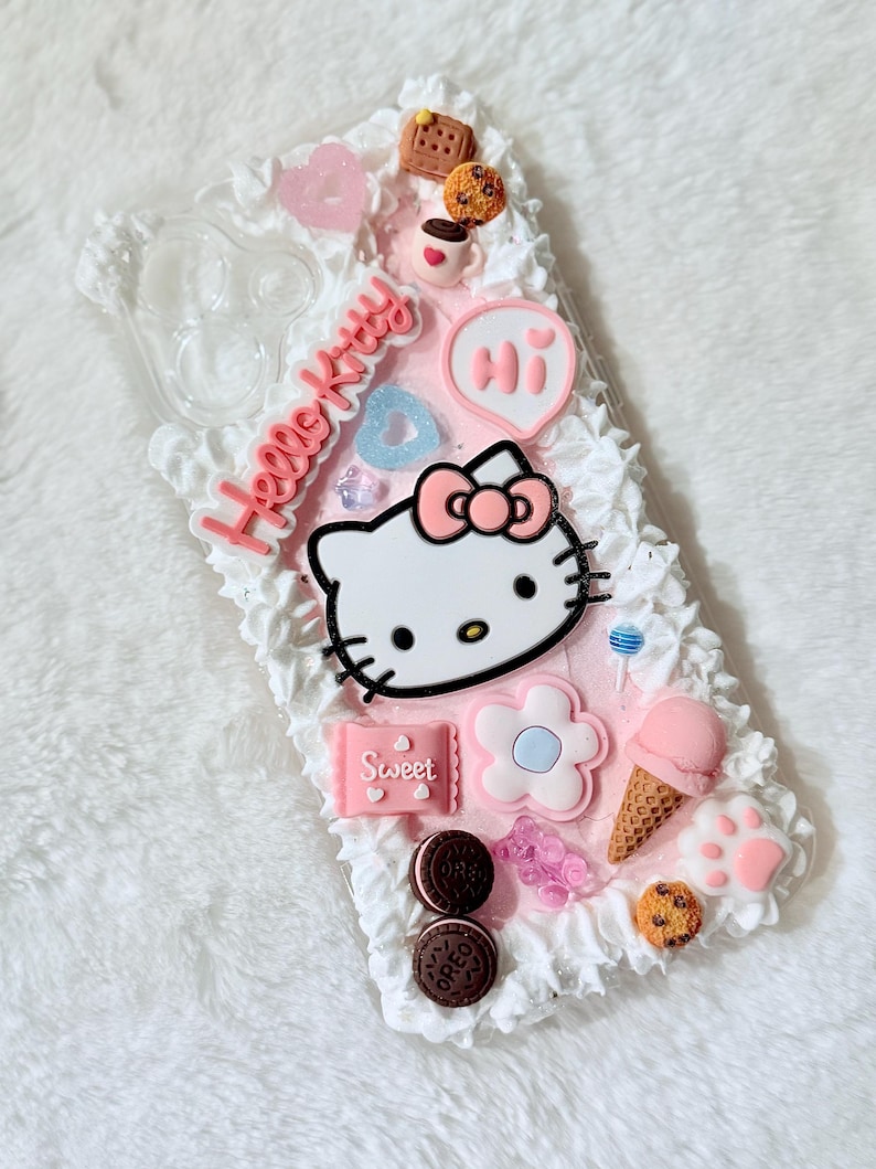 Pu&ograve; includere: Una custodia per telefono rosa e bianca Hello Kitty con un design Hello Kitty 3D, un cuore rosa, una tazza di caff&egrave;, un biscotto, un lecca-lecca, un fiore, un cono gelato e la parola "Ciao" in un fumetto rosa.