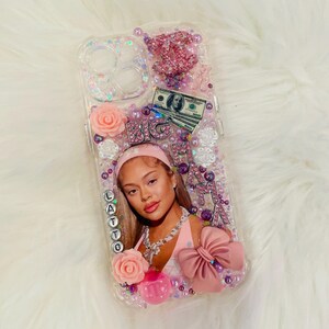 Op de afbeelding: Een transparante telefoonhoes met een roze en wit ontwerp. De hoes heeft een afbeelding van een vrouw met de tekst "BIG LATTO" in roze en witte letters. De hoes is versierd met glitter, strass en bloemen.