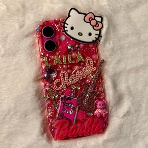 May include: A pink and gold glitter phone case featuring a Hello Kitty character, a pink guitar, a pink book with the words "Burn Book" on it, and the word "Barbie" at the bottom. The case is decorated with pink and gold rhinestones and pearls.