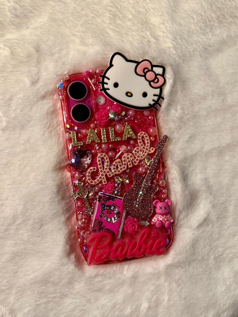 May include: A pink and gold glitter phone case featuring a Hello Kitty character, a pink guitar, a pink book with the words "Burn Book" on it, and the word "Barbie" at the bottom. The case is decorated with pink and gold rhinestones and pearls.