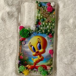 Op de afbeelding: Transparante telefoonhoes met een cartoonafbeelding van Tweety. De hoes is versierd met groene en witte kralen, roze en witte rozen en gouden letters. De hoes is ontworpen om de telefoon te beschermen.