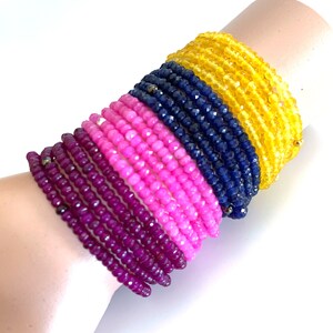 May include: A stack of colorful beaded bracelets in shades of yellow, blue, pink, and purple. The bracelets are made of small, faceted beads and are stacked on a person's wrist.