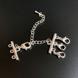 May include: Silver-toned jewelry clasp with a chain and three lobster claw clasps. The clasp has a bar with multiple loops on one end and a hook on the other end.