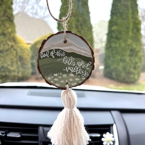 May include: A wooden round ornament with a white tassel hanging from it. The ornament has a painted landscape with the words "God of the hills and valleys" written on it.