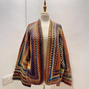 May include: A multi-coloured, open-front crochet cardigan in shades of orange, blue, green, and brown. The handmade garment is displayed on a mannequin, showcasing its open weave and unique colour pattern. This cosy, bohemian-style sweater is perfect for layering.