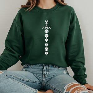 Puede incluir: Una sudadera verde oscuro con un diseño blanco de una espada hecha de dados. Los dados están numerados del 20 al 4.
