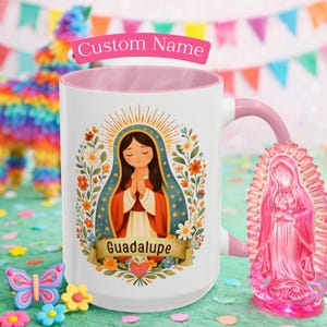 May include: White ceramic mug with a pink interior and handle, featuring a religious illustration of a woman with a halo, surrounded by flowers. The mug has the word "Guadalupe" on a banner. A pink Virgin Mary statue is next to the mug.