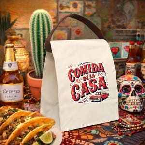 May include: Off-white canvas lunch bag with a brown handle. The bag features the text "Comida de la Casa" in red and blue, with a vintage car graphic. The background includes tacos, beer, and a decorative skull.