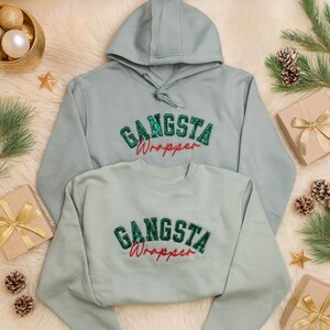 Puede incluir: Dos sudaderas azul claro, una con capucha y otra de cuello redondo, con el texto "GANSTA Wrapper" en verde y rojo. Las sudaderas se muestran sobre una superficie blanca y esponjosa con adornos navideños.